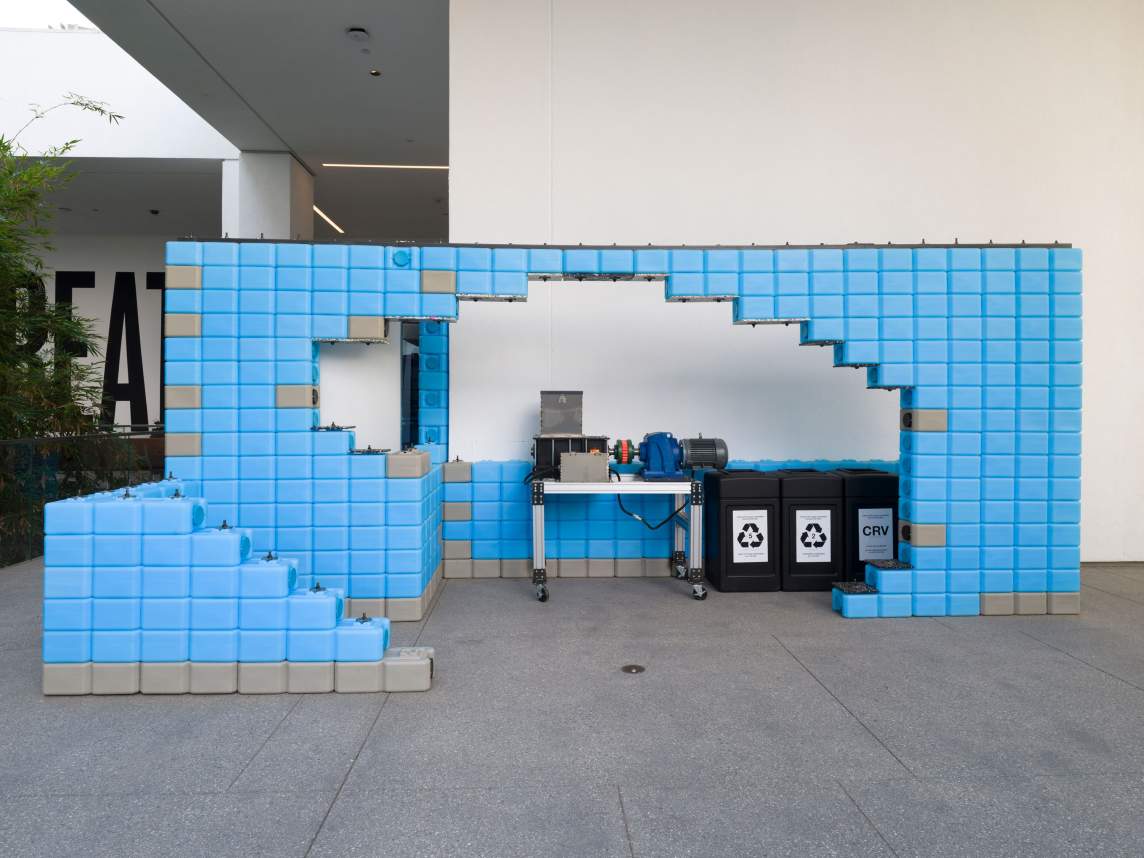 Over Your Head and Under the Weather is an installation formed of a wall with a rounded opening and a side stair composed of blue and gray plastic modular brick containers. Inside the wall is a metal rolling table with an industrial shredder, flanked by a row of black containers with recycling signs.