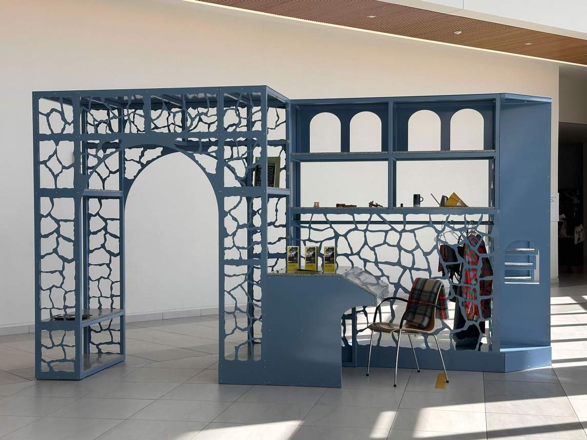 Matters of Consequence is set up in an atrium space lit by windows to our right. A chair is pulled up to the desk, which holds three tabletop signs. A plaid blanket is thrown over the back of the chair, and a few coats hang on the rod on the far side of the structure.