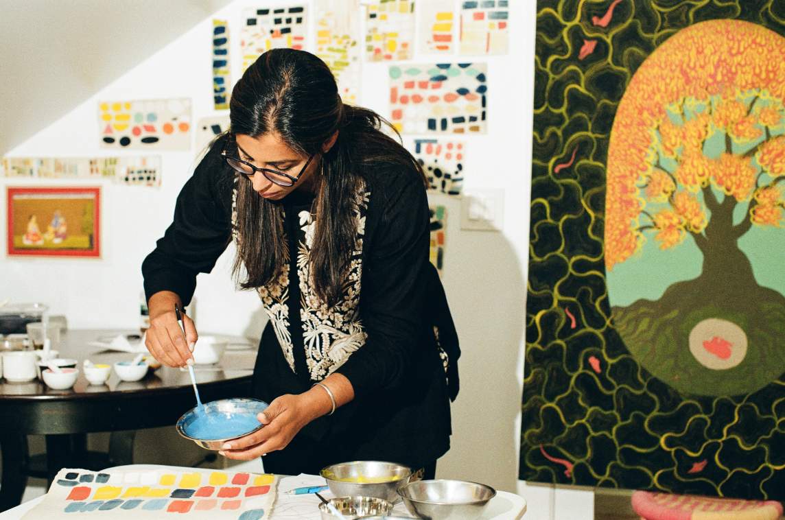 Rupy C. Tut mixes blue paint in a bowl at a table, surrounded by paint samples, with colorful artwork and color swatches displayed on the walls behind her. She is an Indian-American woman with medium skin tone, long dark hair, wearing glasses and a silver bracelet.