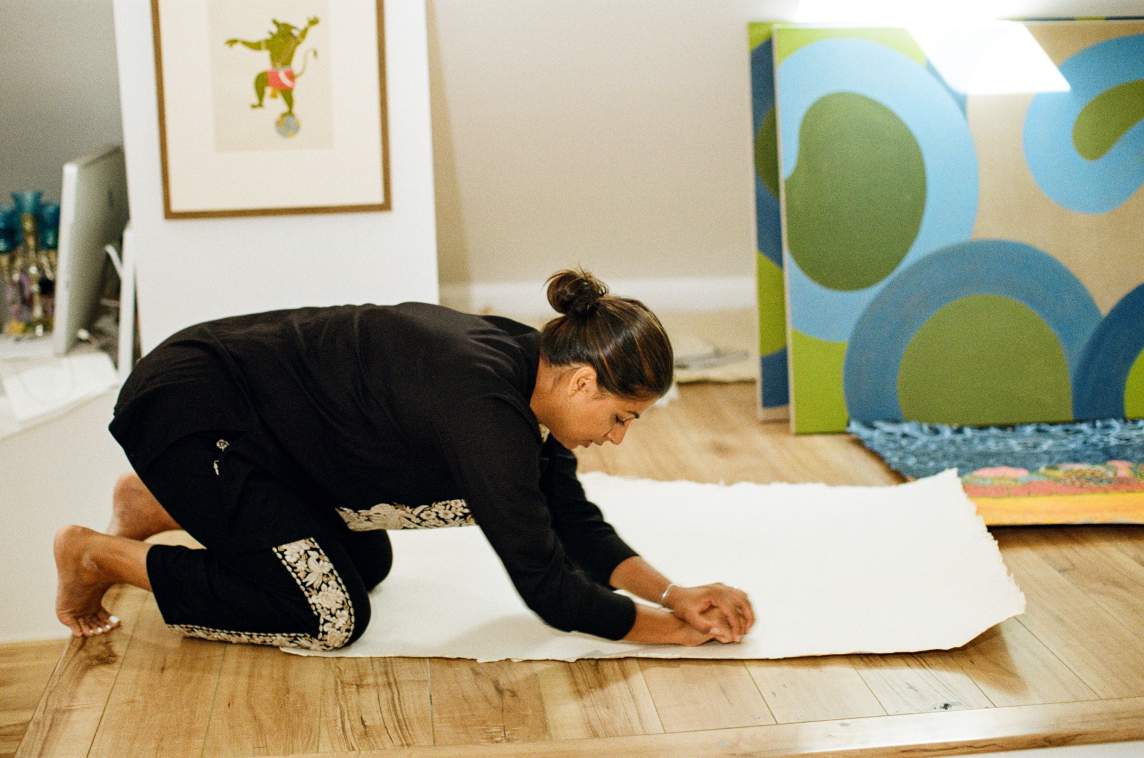 Ruby C. Tut kneels on a wooden floor, pressing her palms into a large sheet of white paper. Colorful artwork and framed pictures are visible in the background. She is wearing a matching black and floral set, and is an Indian-American woman with medium skin tone and long dark hair pulled into a bun.