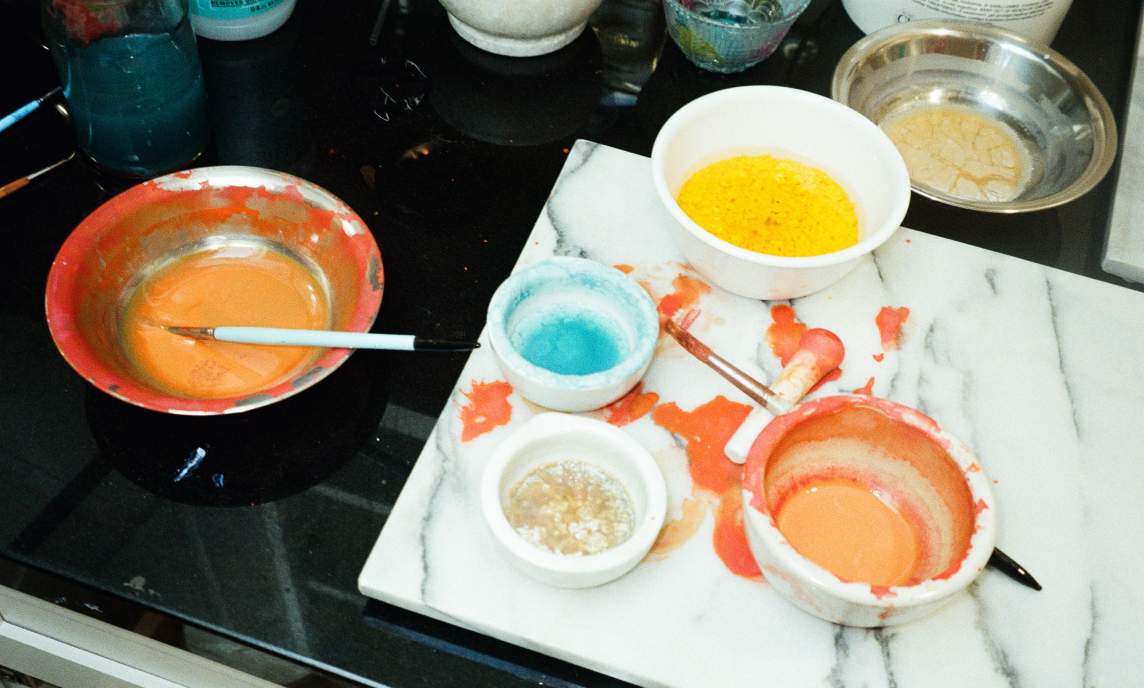 Several bowls containing colorful pigments and liquids, including yellow, blue, and orange, sit on a marble slab and black countertop with paint splatters and mixing tools.