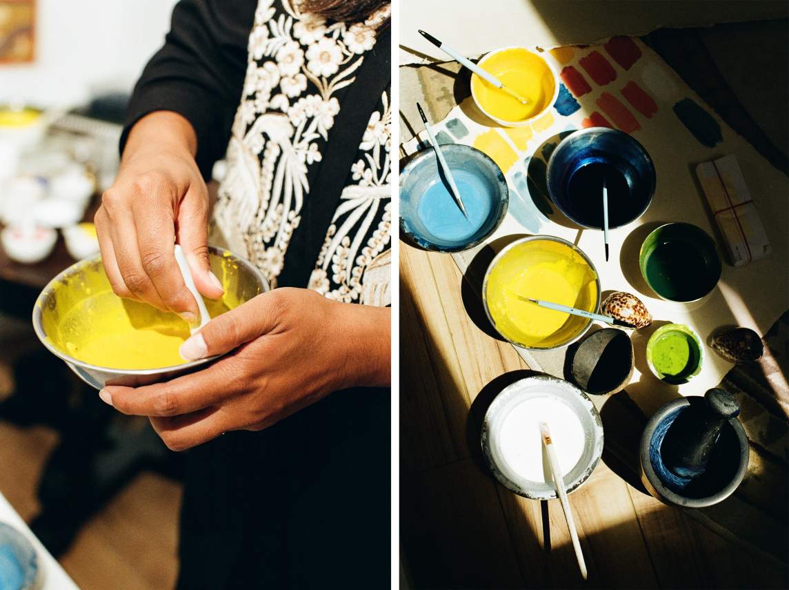 On left, hands mix yellow pigment in a metal bowl. On right, in slanting light, an array of metal bowls hold watery pigments in yellow, blue and green, with a paper with color swatches below.