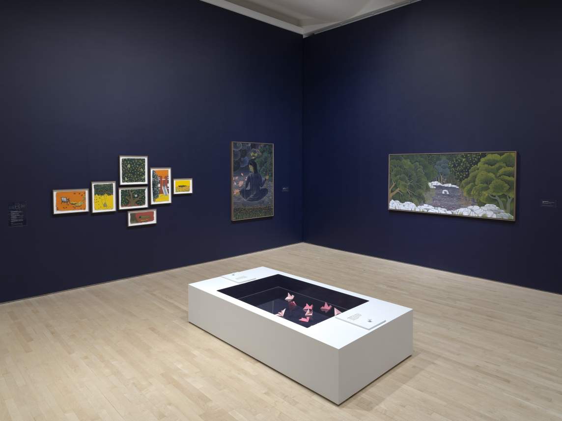 Art gallery with framed paintings on dark walls and a white rectangular display case in the center containing folded pink paper sculptures.