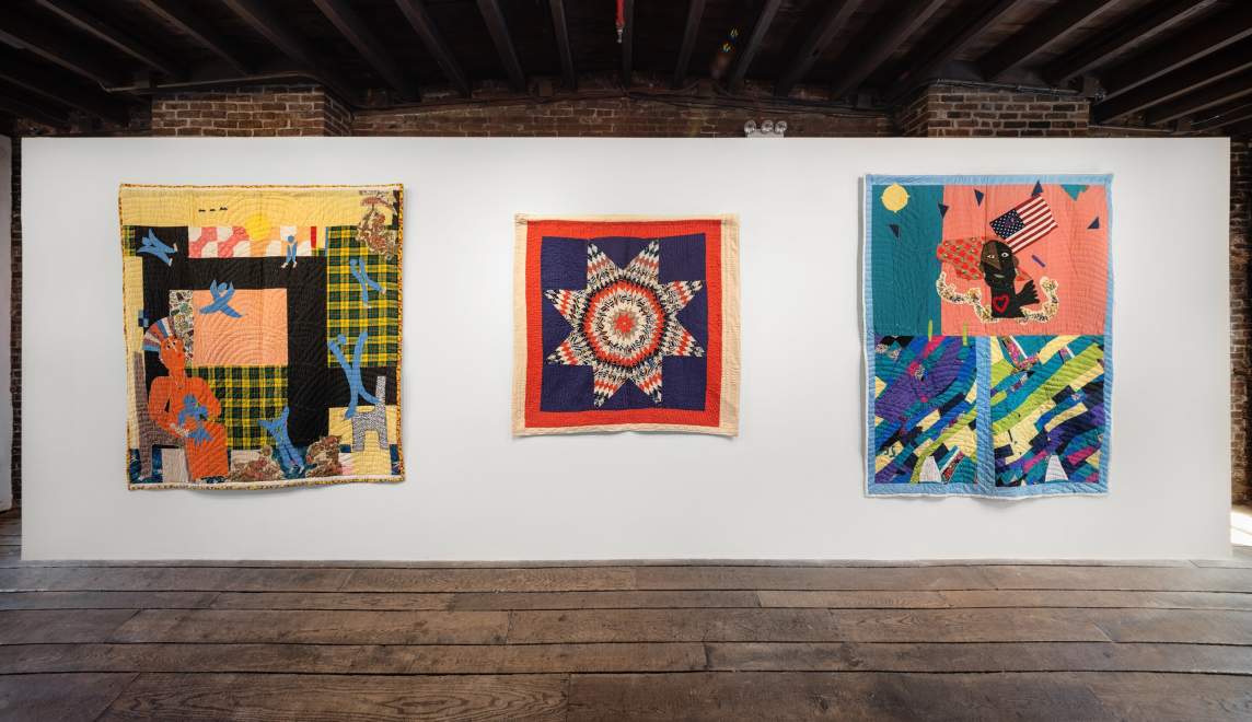 In an art gallery with white walls, distressed wooden floors, and exposed beam ceiling, three quilts hang on the wall. they are brightly colored and contain both geometric and narrative elements.