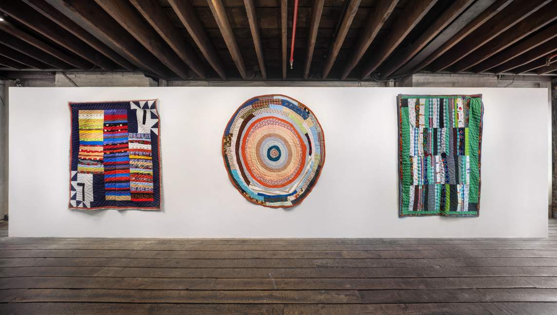 In an art gallery with white walls, distressed wooden floors, and exposed beam ceiling, three quilts hang on the wall. The two outer quilts are rectangular and the central one is circular. All are created by layering fabric strips together to create a composition.