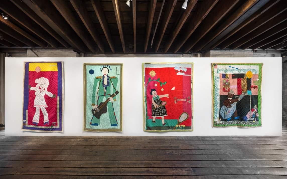 In an art gallery with white walls, distressed wooden floors, and exposed beam ceiling, four rectangular quilts hang on a wall. Each quilt has a central figure depicted either playing a musical instrument or playing a game with paddles.