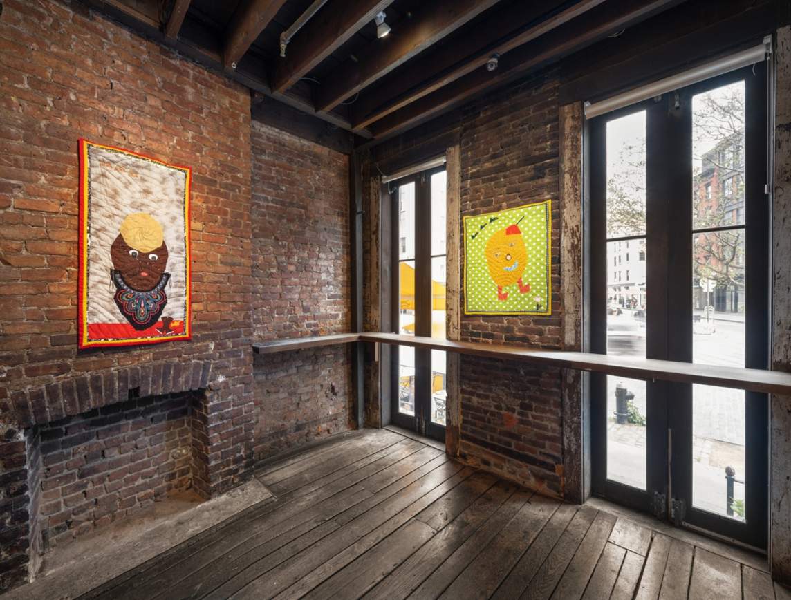 In an art gallery with brick walls, distressed wooden floors, and exposed beam ceiling, two small artworks hang amidst windows. They each depict simplified comical heads.