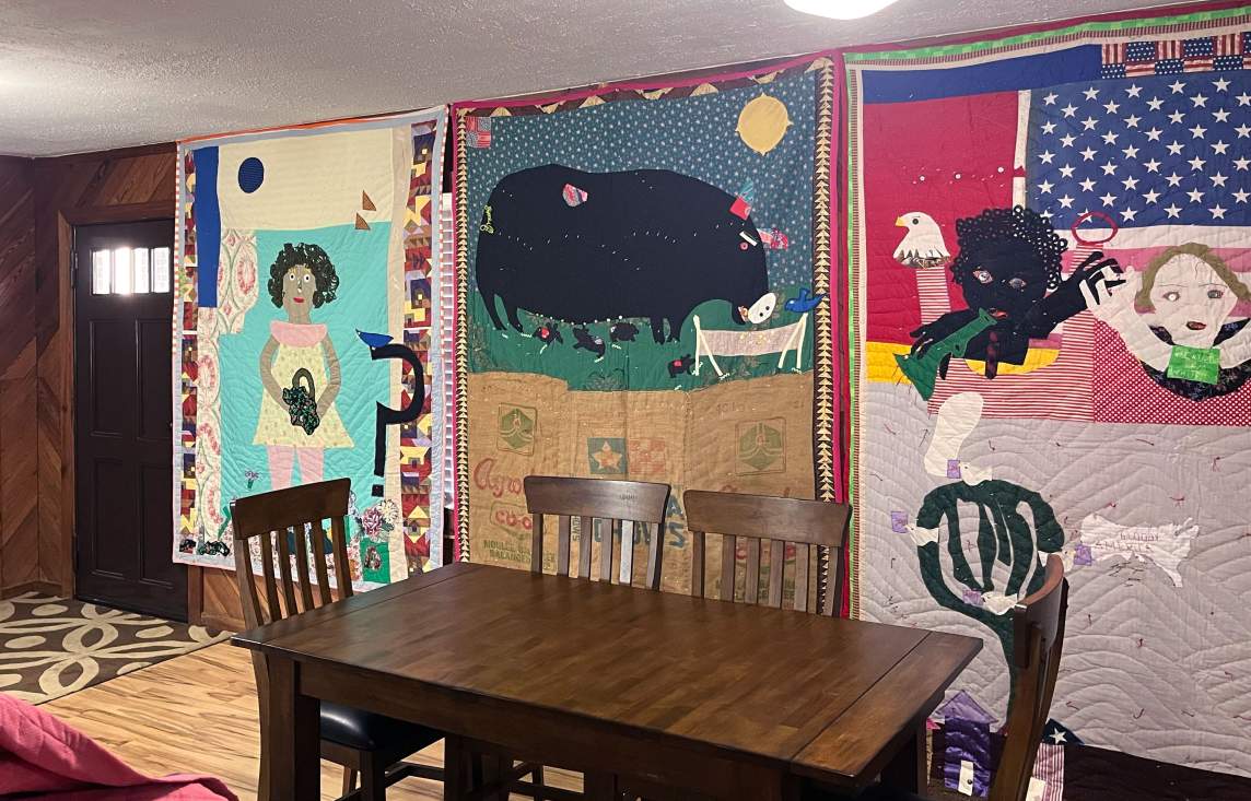 In a dining room with wood paneled walls and a central table, three large quilts with narrative scenes hang on the walls.