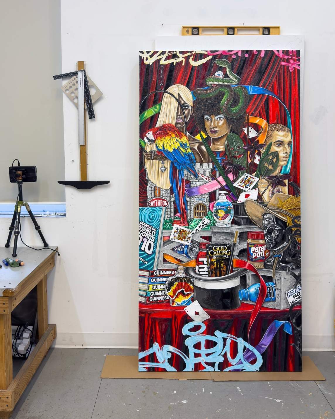 Leaning against a wall in a workspace, a tall rectangular panel is more than twice the height of a table nearby. The painting is filled with Guiness Books of World Records, a jar of Peter Pan peanut butter, a DVD of Good Calling, a bottle of Softsoap, busts, a snake, parrot, and a cinder block against a ruby-red curtain. Graffiti-like letters partially tag the upper and lower edges.