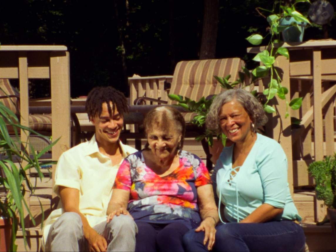 In a film still with warm, saturated color, Sandy Williams IV sits on porch steps with his grandmother and mother, two Black women of mature age with medium skin tone. They wear casual clothes and smile at each other and the camera.