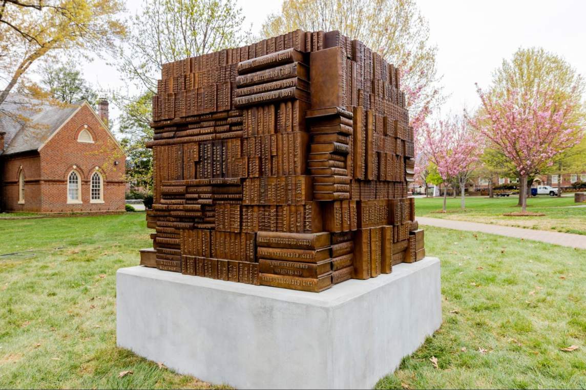 Authors and Architects is a bronze sculpture installed outdoors on what appears to be the grassy lawn of a college campus. The sculpture is a mass of bronze composed of stacks of books that alternate directions, resting on a concrete base.