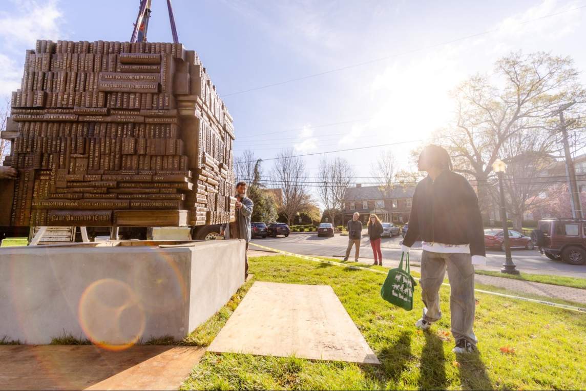 Sandy Williams looks over to a blocky bronze sculpture being lowered onto a concrete plinth. Sun streams down from a blue sky, and brick buildings and trees line the horizon. Sandy is a Black person with medium-light skin tone and chin-length dreadlocks, and carries a totebag.