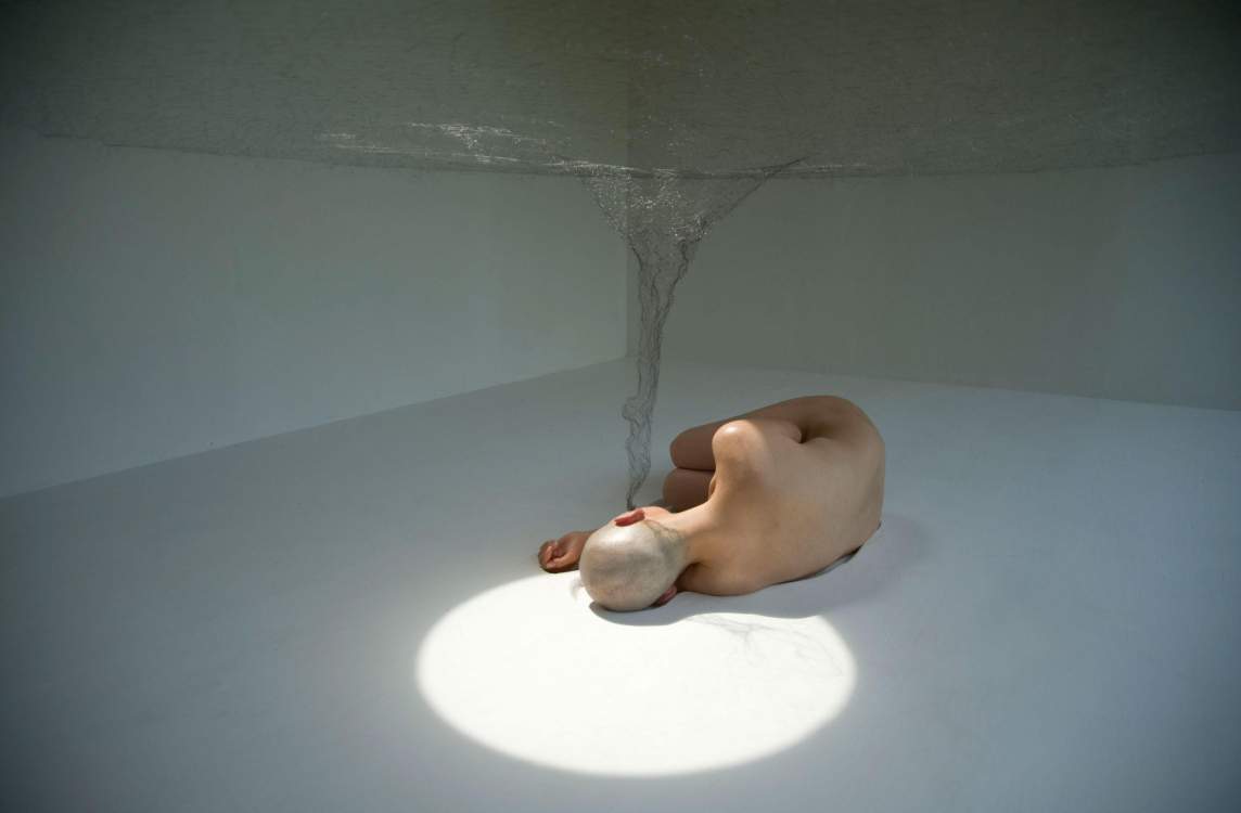 In a photo documenting a performance by artist Jayoung Yoon, a nude figure with light skin tone and a shaved head lays on her side, back to the camera, in the center of a room with white walls and white floors. Her head rests in a halo of light, and a translucent woven sculpture spirals up from her ear in a funnel shape, expanding into a flat surface that hovers a couple of feet above her body.