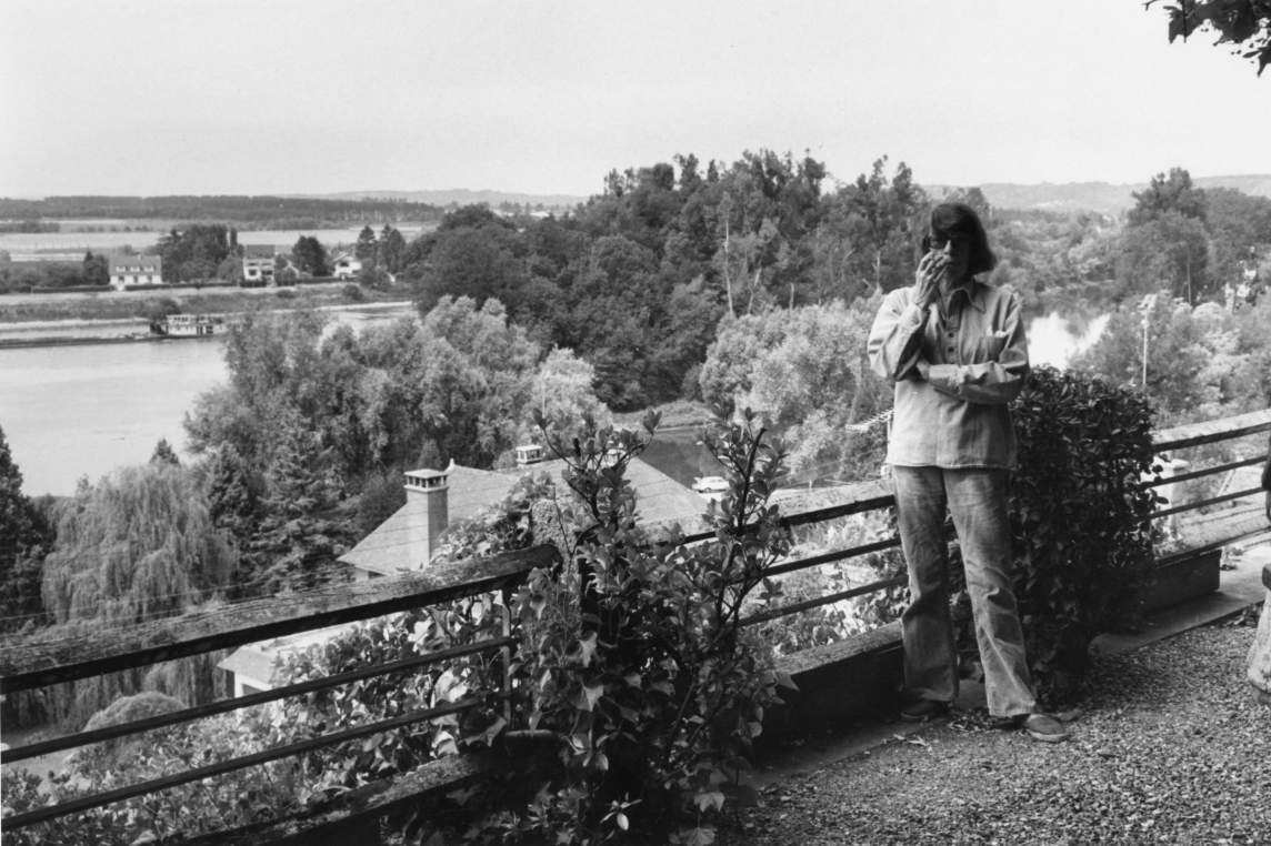 Joan Mitchell stands with folded arms and a cigarette above a sweeping view of the Seine River. She is a white woman in her 60s with dark hair in a bob.