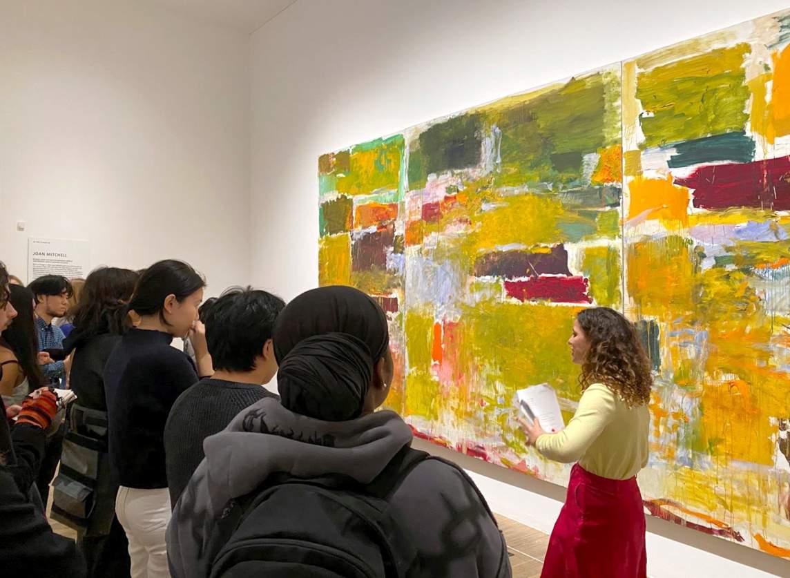 A white woman with curly hair speaks to a group of students in front of a large multi-panel painting, composed of horizontal rectangular fields of greens, ochres, and reds.