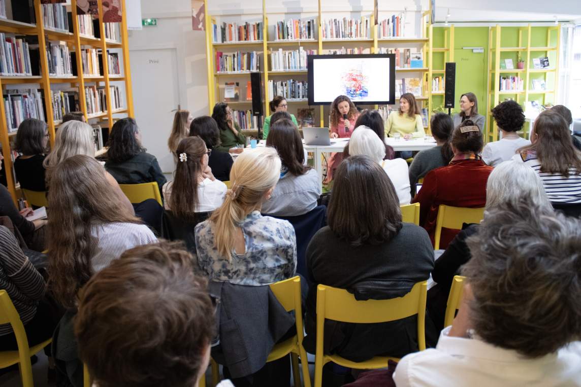 In a room lined with yellow bookshelves, 6 white women sit at a table at the front of the room, as 20+ people in chairs listen. The woman at the center speaks into a microphone.