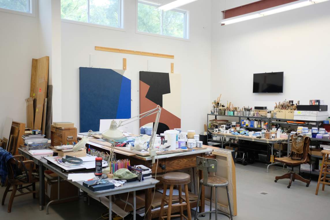 A photo of Virginia Jaramillo's studio in Hamptons, NY, shows a large room with multiple working surfaces, windows up near the ceiling, and paintings resting against the back wall.