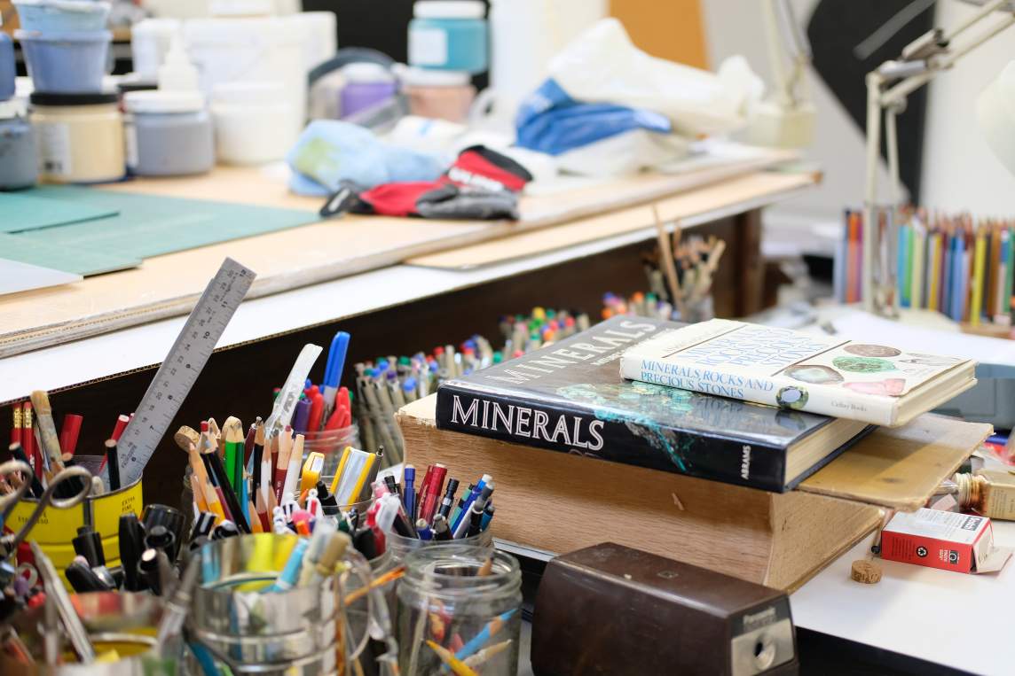 A photo of Virginia Jaramillo's working desk in her studio shows various painting and drawing materials, along with a stack of books on minerals, rocks and semi precious stones.