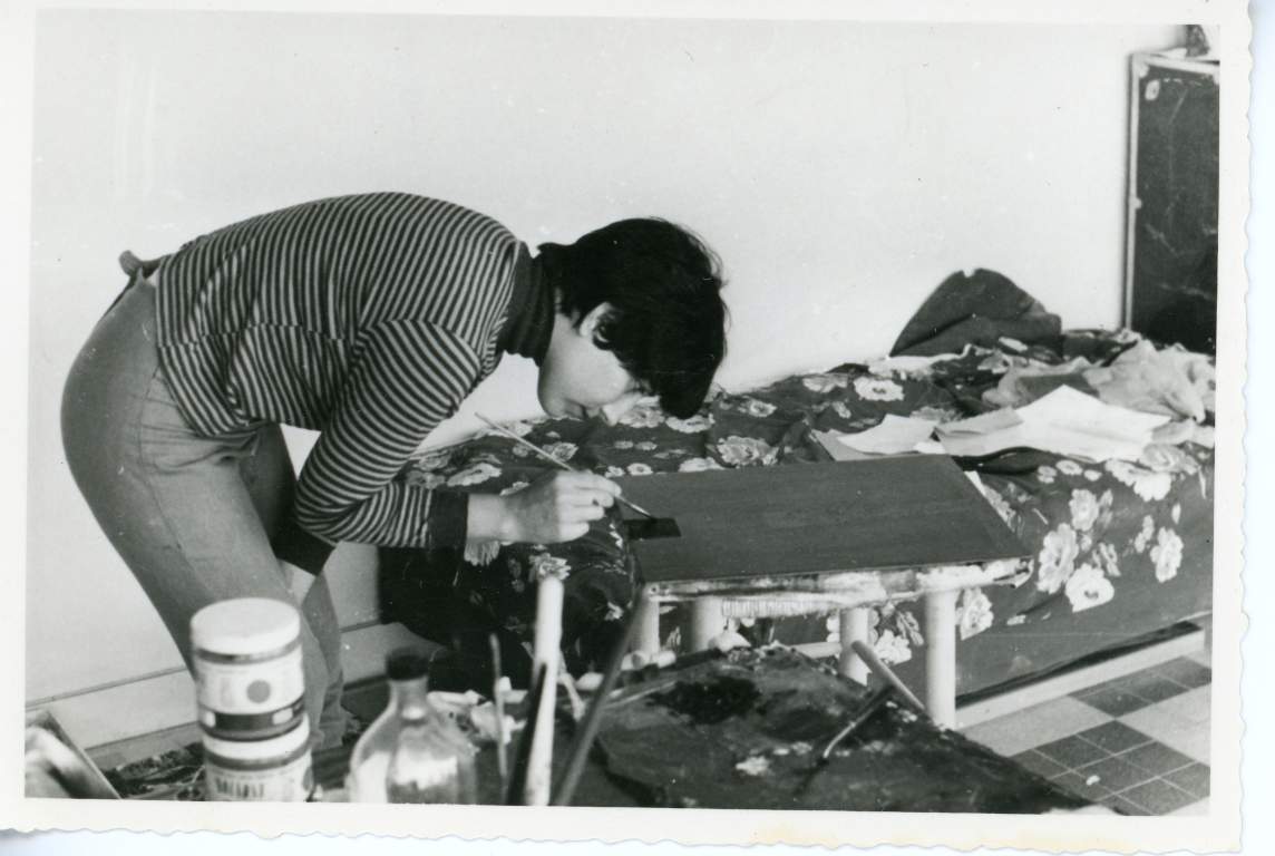 A black and white photo of a woman with short dark hair and light skin, leaning over to paint a dark patch on a small painting that rests on a table.