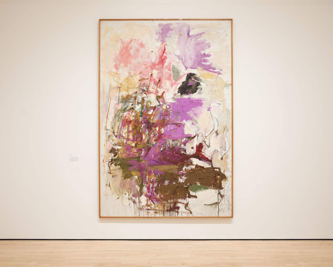 In a gallery space with a wooden floor hangs a large vertical abstract painting by Joan Mitchell. The work has a central form composed of energetic brushstrokes and patches in purple, pink, and brown on a cream ground.