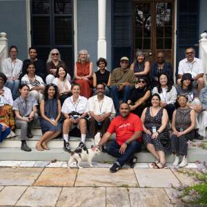 A group of 24 people of various dress, age, and appearance gather on the steps of a Creole cottage style porch, along with a white cat with tabby spots.