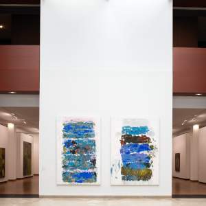 Two abstract paintings hang on a white museum wall in an atrium with wood paneled architecture and galleries seen behind the central wall. The paintings are vertical in orientation and each feature broad bands of horizontal brushstrokes predominantly in vibrant blue and green hues.
