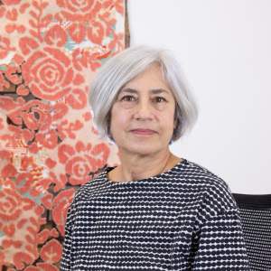 Elana Herzog, a white woman with short gray hair sits on a black chair, wearing a black and white patterned shirt, with a red floral textile artwork in the background.