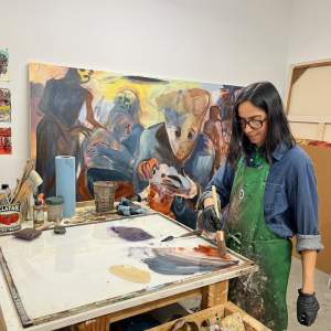 ala Iqbal, a Pakistani-German-American woman with medium-light skin tone, shoulder-length dark hair, and black glasses, mixes paint on a glass panel in her painting studio. On the walls behind her we see paintings big and small and a group of stacked canvases.