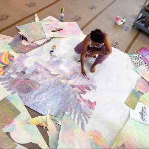 Image is an aerial view of the artist working on a large studio floor. She squats barefoot on a massive drawing collagescape of a post-apocalyptic island battle where three ships hover above a city and spirits are resurrected for an impending battle. The drawing is surrounded by sheets of candy colored prints and watercolors, and is constructed using hyper-feminized colors and material.