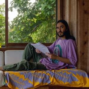 Drew Kahuʻāina Broderick, a native Hawai’ian with medium-light skin tone, long brown hair, and a beard looks towards the camera as he reclines on a window seat holding papers, wearing a purple t-shirt and green jeans. Lush vegetation is visible through the large sliding window.