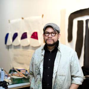 Leslie, a Black man with medium skin tone, stands in his studio amidst a backdrop of artwork and painting materials. He faces the camera, wearing dark glasses, a newsboy cap, a close-trimmed beard, and a painter’s smock over a black button-down shirt.