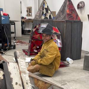 Chiffon Thomas sits on a concrete studio floor surrounded by sculptures and materials, wearing a heavy canvas jacket, gloves and a raised welding mask. He looks towards the camera with a slight smile, and is a Black man with a medium skin tone and dark short hair.