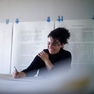 A Black woman with curly dark hair and medium-light skin tone sits at a table with pencil in hand, smiling and looking down at her work. Behind her, oversized typed pages are taped to the wall.