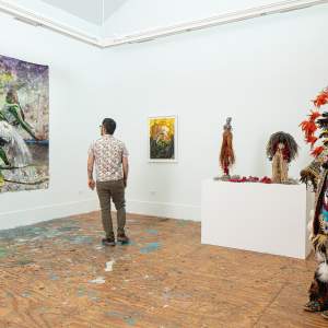 A man stands in a gallery space looking at a large colorful work on paper of two people dancing and bumping pelvises. To his right is a smaller abstract work, two small figurative mixed media sculptures, and a full-size beaded costume with feathered regalia.