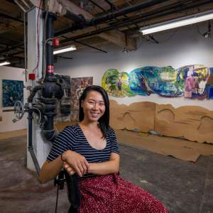 Kathy Liao sits in the foreground of an industrial artist’s studio space, with artworks and tools visible on the walls behind her. She is a Taiwanese-American woman with dark hair. She wears a striped blue-and-white top and a red skirt with a speckled pattern. Her arms rest on the back of the chair and in front of her chest, and she smiles at the camera.