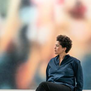 Julie Mehretu sits in profile in front of a large, blurred abstract painting. She short, dark, curly hair, a medium light skin tone, and wears a navy button up shirt. A smaller abstract artwork with lines and shapes is visible on the wall.