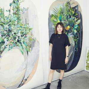 Naomi Kawanishi Reis stands in front of two large oval abstracted botanical paintings. She is a Japanese-American woman, wearing a black dress, and black boos, with mid length dark hair.