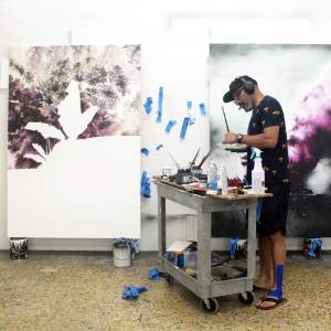 Gamaliel Rodríguez mixes paint in front of colorful paintings in progress of clouds and palm trees in his studio. He is a Puerto Rican man with medium light skin tone, graying beard and wears glasses, a black baseball hat, headphones, and black shirt and shorts.