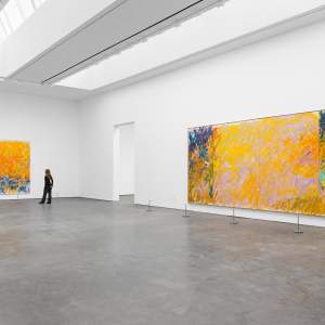 A person views two large abstract paintings by Joan Mitchell with dominant yellow and orange tones in a spacious, modern art gallery with white walls and a concrete floor. The painting on the left is a large diptych, and on the right is a quadriptych.