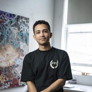 Anthony White leans on a saw horse in a studio in front of a painting and a window. He is a multi-racial person with medium skin tone, short curly dark hair and wears all black.