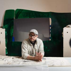 Leslie Smith sits at a table layered with canvas, in front of abstract panels, and two large speakers. He is a Black man with medium light skin tone and dark beard, and wears glasses, a cap, and a paint-splattered button-up.