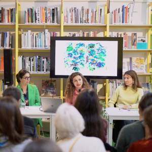 5 white women of various dress sit at a table in a library in front of a screen with an abstract painting displayed.