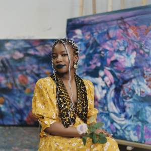 Jadé Fadojutimi looks directly at the camera serenely, holding a rose in her lap. She is a Black woman with medium skintone, long braided hair plaited with black and white strands. She wears black lipstick and a yellow floral dress, behind her are two large abstract paintings.