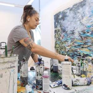 Artist Ann Marie Auricchio in a grey shirt mixes paint on a table with splatted acrylic paint bottles in a bright studio with a large abstract painting in the background