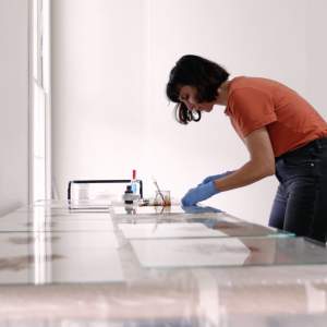 Artist Manon Bellet leans over a table covered with panes of glass, applying dark liquid