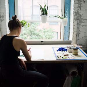 Anne Buckwalter, a white woman with brown hair in a bun in a black tank top, faces away and paints at a drafting desk in a studio in front of a window.
