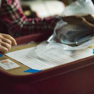 Hands with light skin hold a ceramic item above a cardboard box with taped paper inventory on top.