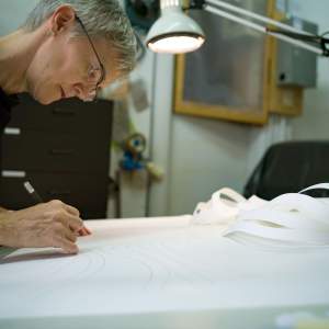 Heather Cox, a woman with light skin, short cropped gray hair and wire rim glasses, leans over and cuts undulating lines from a large sheet of white paper with an x-acto knife.