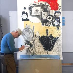 Marcos Dimas, a Puerto Rican man with medium tone skin, short gray hair and glasses, takes notes on a legal pad in front of a large artwork on paper.