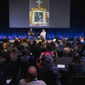 Josh T. Franco and Amalia Mesa-Bains sit on a stage in a room filled with people sitting at round black tables. A screen behind them shows one of Amalia's artworks.